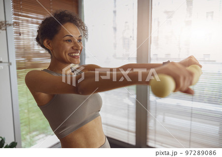 Smiling woman with dumbbells doing exercising at home and working on arms strength Smiling woman with dumbbells doing exercising at home and working on arms strength 97289086