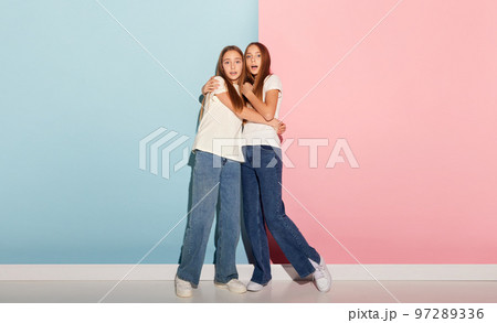 School age girls, sisters friends wearing white t-shirts and jeans communicate isolated over pink-blue background. Concept of love, friendship, family, education, hobbies 97289336