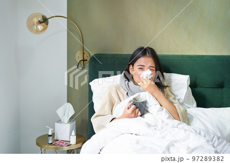 Covid-19, flu and vaccination concept. Korean girl lying in bed with cold, catching influenza, drinking hot tea and using prescribed medication 97289382