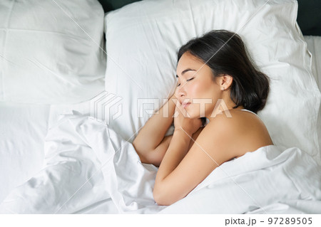 Top view image of asian woman sleeping alone in king size bed on white pillows. Young girl lying in her bedroom with eyes closed, morning sunlight shines in room Top view image of asian woman sleeping alone in king size bed on white pillows. Young girl lying in her bedroom with eyes closed, morning sunlight shines in room 97289505