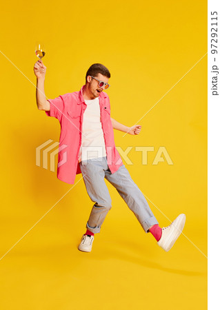 Portrait of young man in pink shirt dancing with wine glass isolated over vivid yellow background. Birthday party Portrait of young man in pink shirt dancing with wine glass isolated over vivid yellow background. Birthday party 97292115