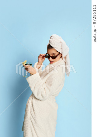 Portrait of beautiful girl in bathrobe and sunglasses, drinking cocktail over blue background. After shower. Relaxation. Party 97295111