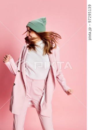 Portrait of young beautiful girl in a suit, hat posing, dancing over pink background. Good mood, positive vibe 97295139