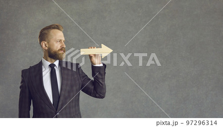 Serious businessman holding arrow and looking straight ahead on grey copy space background Serious businessman holding arrow and looking straight ahead on grey copy space background 97296314