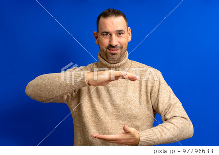 Happy hispanic man wearing a brown turtleneck showing something between his hands, product marketing advertisement concept with copy space over blue background. Happy hispanic man wearing a brown turtleneck showing something between his hands, product marketing advertisement concept with copy space over blue background. 97296633