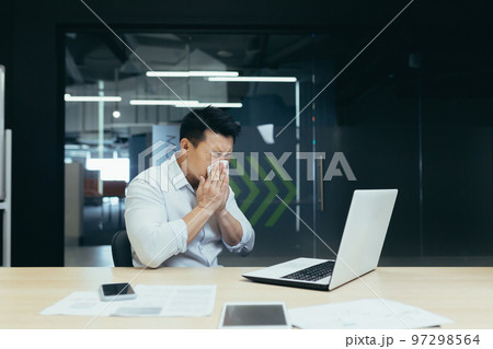Illness at work. A young Asian man sits in the office at the table, wipes his nose with a napkin, has a runny nose, flu, feels bad, is exhausted. Illness at work. A young Asian man sits in the office at the table, wipes his nose with a napkin, has a runny nose, flu, feels bad, is exhausted. 97298564