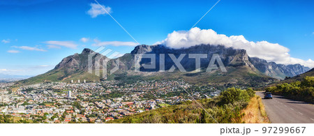 A mountain road overlooking the city with a cloudy blue sky. Panoramic landscape of green mountains surrounding an urban town and a scenic road for traveling along Cape Town, South Africa A mountain road overlooking the city with a cloudy blue sky. Panoramic landscape of green mountains surrounding an urban town and a scenic road for traveling along Cape Town, South Africa 97299667