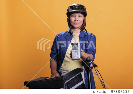 Portrait of deliverywoman with blue uniform and helmet delivering fast food order giving POS terminal to customer. Pizzeria employee using bike as transportation while carrying takeout food backpack Portrait of deliverywoman with blue uniform and helmet delivering fast food order giving POS terminal to customer. Pizzeria employee using bike as transportation while carrying takeout food backpack 97307157
