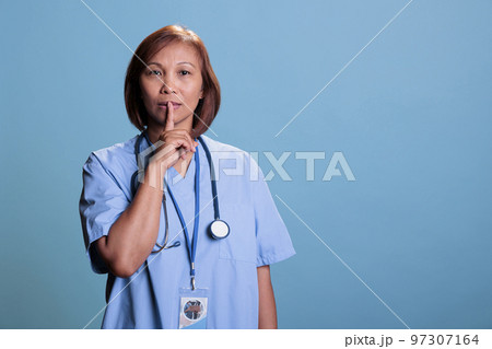 Senior nurse showing shhh taboo sign with finger to lips while working at medical expertise during checkup visit appointment. Specialist assistant wearing blue uniform and stethoscope. Senior nurse showing shhh taboo sign with finger to lips while working at medical expertise during checkup visit appointment. Specialist assistant wearing blue uniform and stethoscope. 97307164