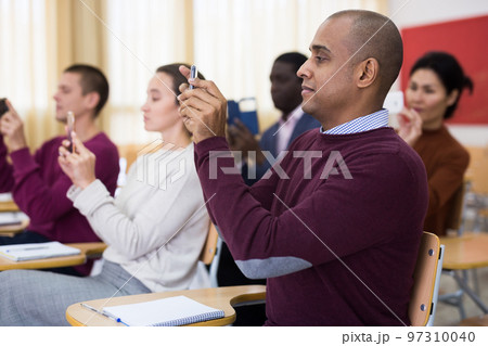 Students of different age sitting together at tables, shooting lesson on smartphones in classroom 97310040