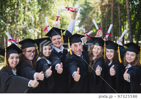 A group of graduates in robes give a thumbs up outdoors. Elderly student. 97325586