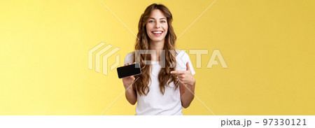 Carefree charismatic cute curly-haired female gamer likes playing smartphone games showing own game score hold mobile phone horizontal pointing display laughing pleased amused 97330121