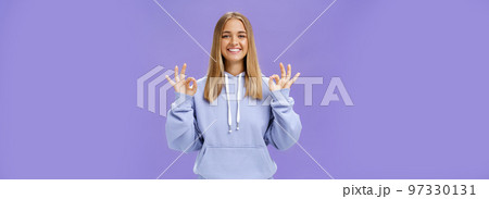 Girl got everything under control. Delighted happy charming woman with fair hair and tanned skin without makeup showing okay gesture smiling assured and pleased posing in hoodie over purple wall Girl got everything under control. Delighted happy charming woman with fair hair and tanned skin without makeup showing okay gesture smiling assured and pleased posing in hoodie over purple wall 97330131