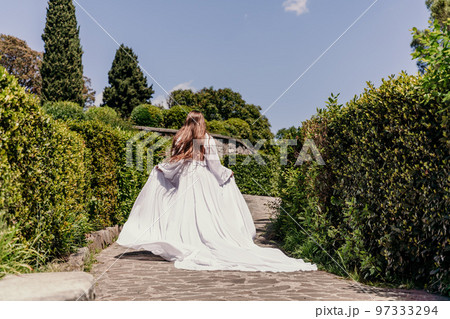 a beautiful woman with long brown hair and long white dress runs along a path along beautiful bushes in the park rear view 97333294