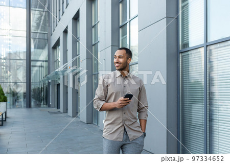 Smiling hispanic man walking down street near modern office building, freelancer businessman looking away holding mobile phone. 97334652