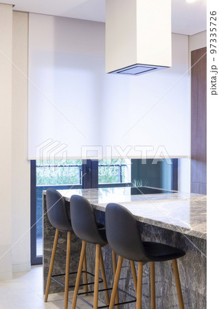 Roller blind large size on the window in the interior. Motorized roller shade white color in the kitchen. Bar counter with built-in hob and hood above it are near the window. Smart home concept.  Roller blind large size on the window in the interior. Motorized roller shade white color in the kitchen. Bar counter with built-in hob and hood above it are near the window. Smart home concept.  97335726