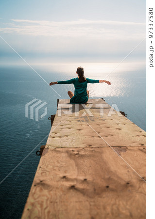 A girl sits on a wooden springboard for jumping with a rope. In a dark green dress with her hands in the air. A girl sits on a wooden springboard for jumping with a rope. In a dark green dress with her hands in the air. 97337580