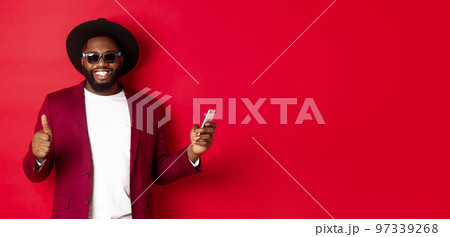 Handsome african american man using phone app and showing thumb up, smiling at camera, wearing sunglasses and fancy hat, red background 97339268