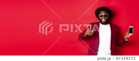Handsome and stylish Black man showing phone screen and thumb up at camera, recommending online store app, red background Handsome and stylish Black man showing phone screen and thumb up at camera, recommending online store app, red background 97339273