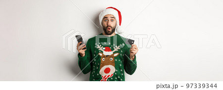 Online shopping and winter holidays concept. Surprised man buying gifts in internet with mobile phone and credit card, standing in Santa hat over white background 97339344