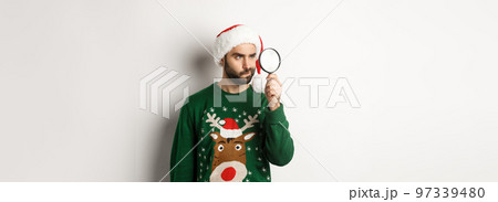 Christmas and holidays concept. Young man in Santa hat searching for something with magnifying glass, standing over white background 97339480