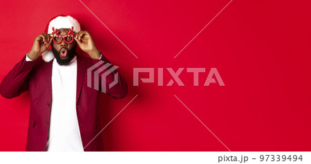 Funny Black man celebrating New Year, wearing party glasses and santa hat, having fun over red background Funny Black man celebrating New Year, wearing party glasses and santa hat, having fun over red background 97339494