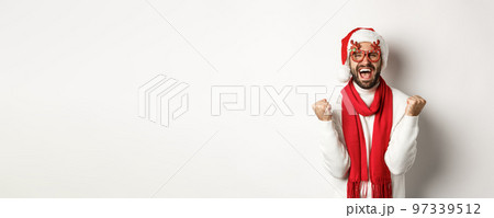 Christmas, New Year and celebration concept. Excited man in Santa hat and party glasses, winning and rejoicing, making fist pump, shouting yes, standing over white background Christmas, New Year and celebration concept. Excited man in Santa hat and party glasses, winning and rejoicing, making fist pump, shouting yes, standing over white background 97339512