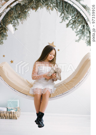 Cute girl holding a soft favorite toy and sitting on a swing decorated for Christmas Cute girl holding a soft favorite toy and sitting on a swing decorated for Christmas 97339916