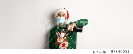 Concept of covid-19 and Christmas holidays. Happy guy in face mask and santa hat, celebrating New Year party and pouring glass of champagne, white background 97340051