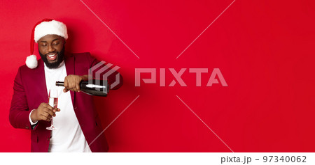 Christmas, party and holidays concept. Handsome Black man in santa hat pouring glass of champagne and smiling, celebrating New Year, red background Christmas, party and holidays concept. Handsome Black man in santa hat pouring glass of champagne and smiling, celebrating New Year, red background 97340062