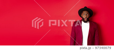 Fashion and party concept. Shocked and displeased black man staring at something unpleasant, looking upper left corner and frowning, standing against red background Fashion and party concept. Shocked and displeased black man staring at something unpleasant, looking upper left corner and frowning, standing against red background 97340079