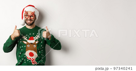 Christmas holidays, celebration concept. Happy man in Santa hat and party glasses showing thumbs up in approval, like something, standing over white background 97340241