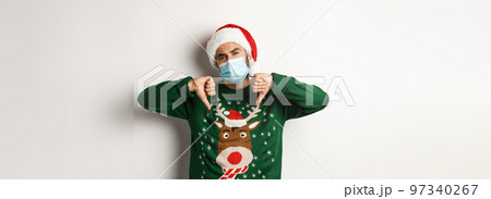 Concept of covid-19 and Christmas holidays. Displeased guy in face mask and santa hat showing thumbs down, standing over white background 97340267