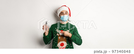 Concept of covid-19 and Christmas holidays. Young man in medical mask offer an antiseptic, pointing at hand sanitizer, wearing Santa hat, white background 97340848