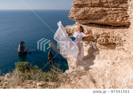 A beautiful young woman in a white light dress with long legs stands on the edge of a cliff above the sea waving a white long dress, against the background of the blue sky and the sea. 97341501