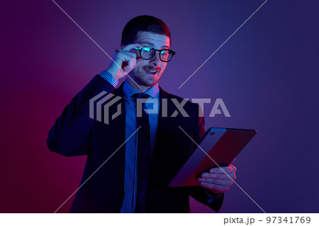 Young man, manager wearing business suit and eyeglasses using digital tablet on gradient blue purple background in neon light. Studying, work, job, finance 97341769