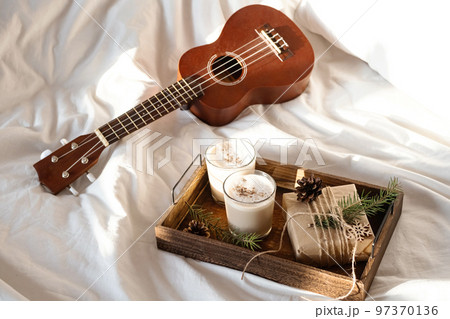 Christmas cozy morning at home. Gift box and eggnog on wooden tray near ukulele guitar on bed. 97370136