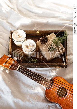 Christmas cozy morning at home. Gift box and eggnog on wooden tray near ukulele guitar on bed. 97370138