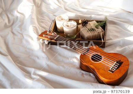 Christmas cozy morning at home. Gift box and eggnog on wooden tray near ukulele guitar on bed. 97370139