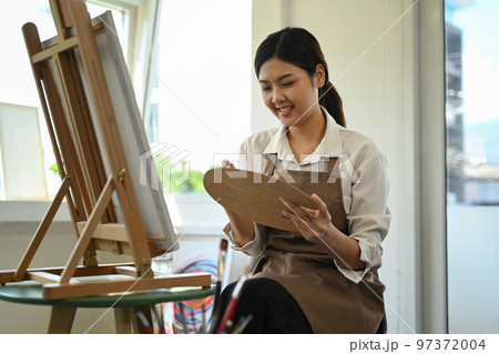 Smiling asian student woman in apron painting in watercolor on easel. Education, hobby, art concept Smiling asian student woman in apron painting in watercolor on easel. Education, hobby, art concept 97372004