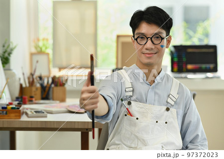Handsome artist man holding paintbrushes and palette smiling to camera while sitting in art studio Handsome artist man holding paintbrushes and palette smiling to camera while sitting in art studio 97372023