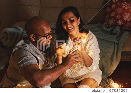 Happy interracial couple celebrating New Year at home together, sitting by Christmas tree Happy interracial couple celebrating New Year at home together, sitting by Christmas tree 97382095