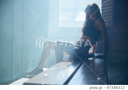 Young girl of an athletic build in the locker room of a sports club wears boxing gloves, a girl is dressed in sports black underwear, sun rays from window, sneakers 97386322