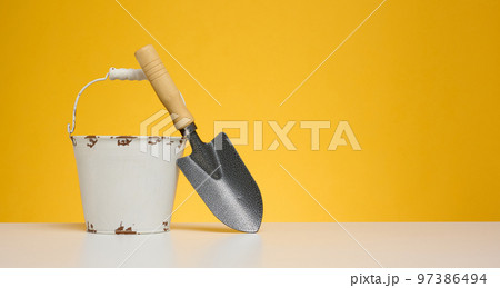 White metal bucket and miniature garden shovel on white table, yellow background White metal bucket and miniature garden shovel on white table, yellow background 97386494