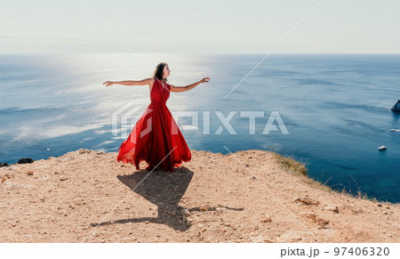 Woman in red dress on sea. Side view a Young beautiful sensual woman in a red long dress posing on a rock high above the sea on sunset. Girl on the nature on blue sky background. Fashion photo. 97406320