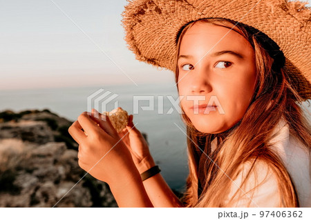 Happy girl eating corn. Summer snacking on the sea. Portrait of young beautiful girl in straw hat eating grilled corn while sitting by the sea on sunset time. Close up. Selective focus 97406362