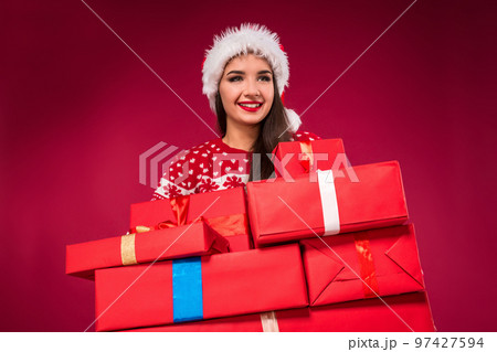 Close-up portrait of amazed beautiful woman holding many presents on red background. Young woman in santa hat looks away 97427594