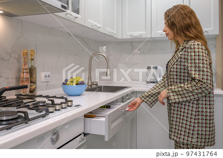 Cute young housewife pulls out the cabinet drawer at kitchen Cute young housewife pulls out the cabinet drawer at kitchen 97431764