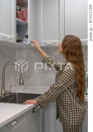 Cute young housewife opens the kitchen cabinet door Cute young housewife opens the kitchen cabinet door 97431766