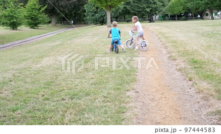Sport activity for children. Small kids playing bicycle and scooter riding on path in park in summer drought dry grass. Little Brother and Sister Siblings playing together. Child development concept 97444583